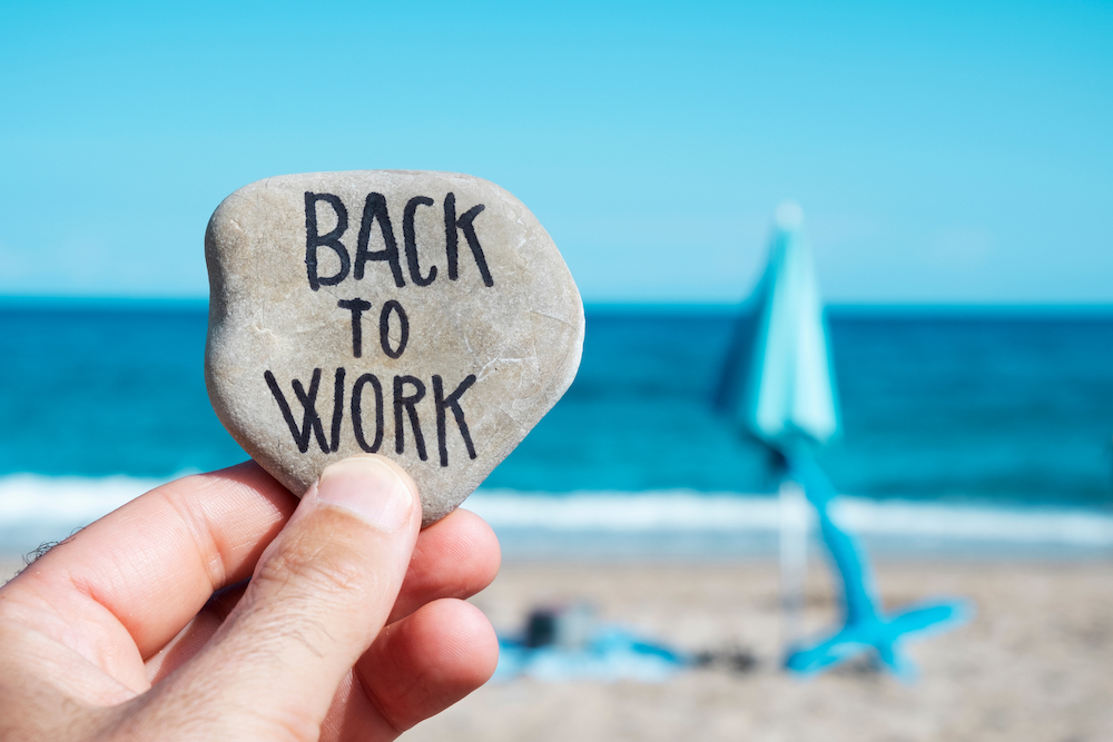 closeup of the hand of a young caucasian man on the beach, in front of a folded beach umbrella and the ocean in the background, holding a stone with the text back to work written in it